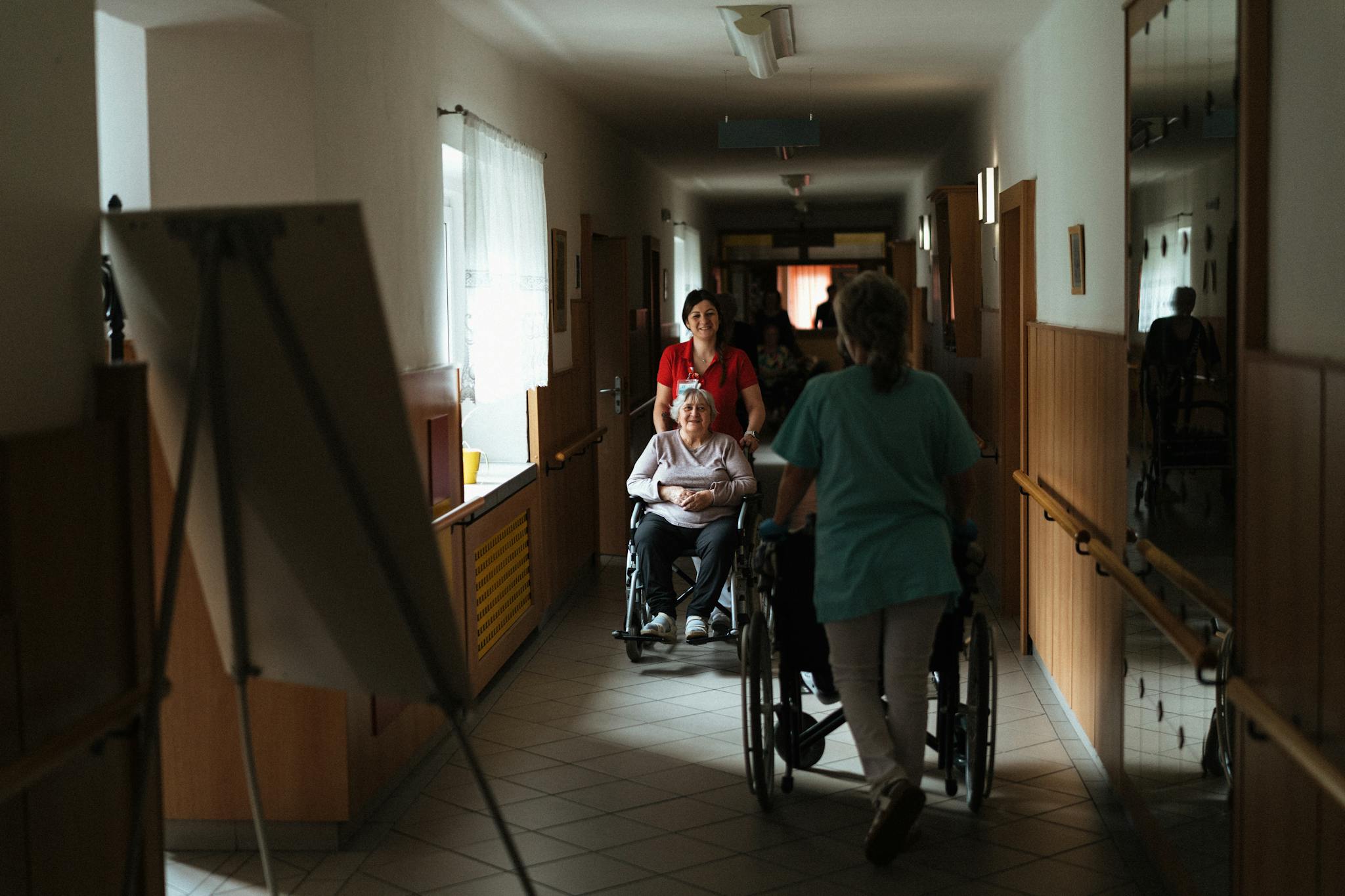 Caregiver assisting elderly woman in a wheelchair through a retirement home corridor.