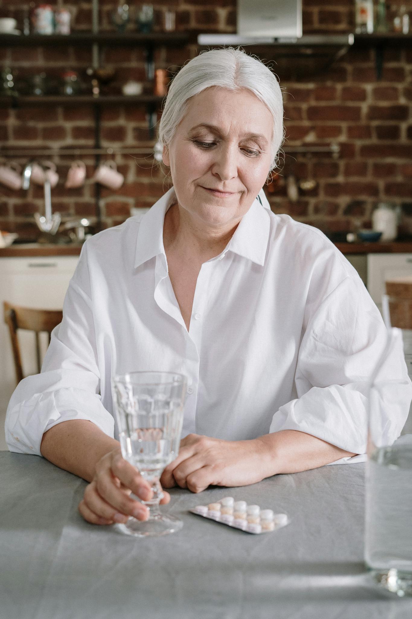 Senior woman in white shirt sitting at dining table with water and medication in a cozy home kitchen.